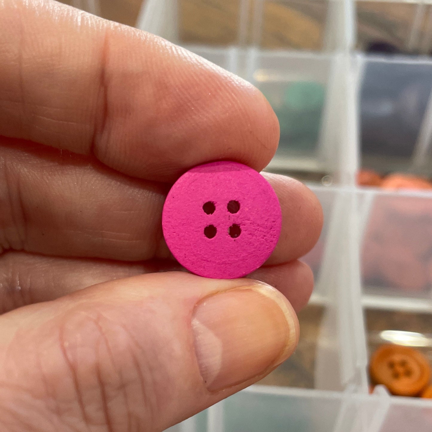 Hand holding a pink button with a container of assorted buttons in the background