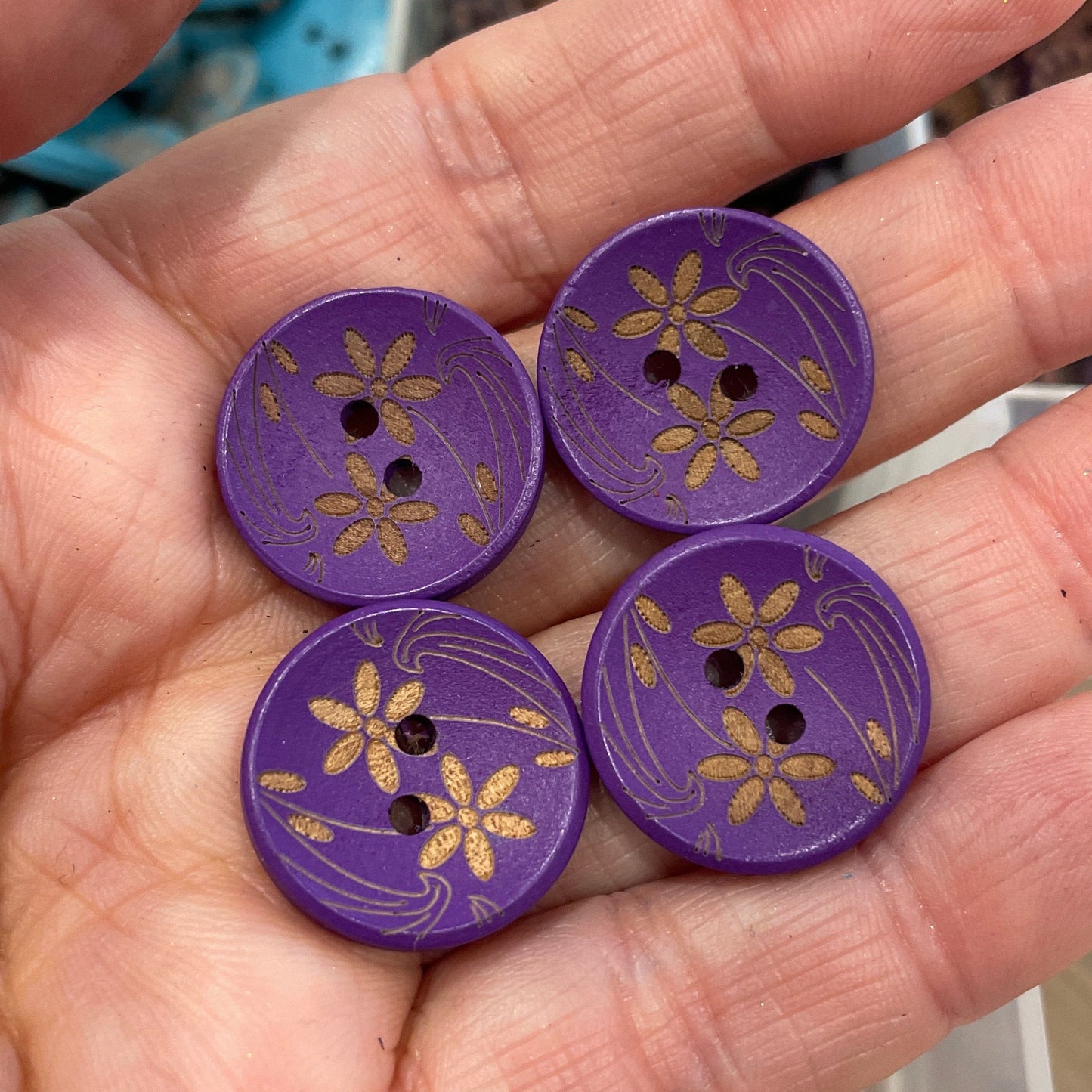 Purple buttons with floral designs held in a hand, with a container of buttons in the background.