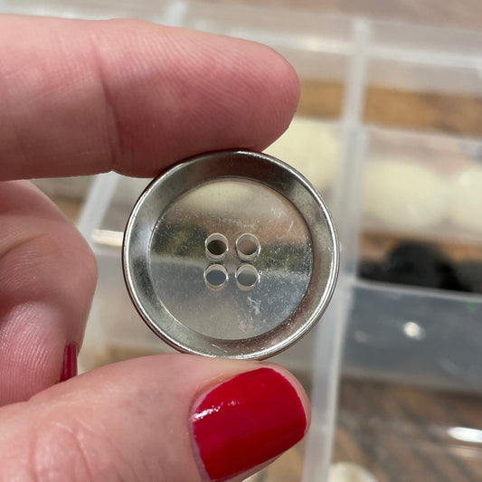 Hand holding a metal button with a clear container of buttons in the background