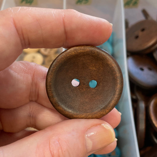 Hand holding a wooden button with a blurred container of buttons in the background