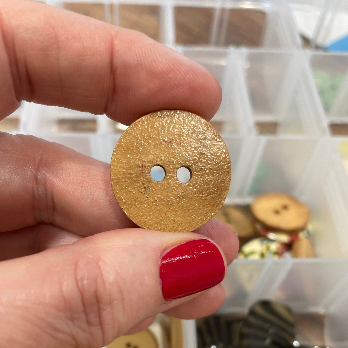 Gold button held by a hand with red nail polish, against a background of button collection.