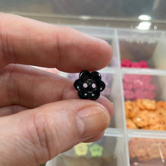 Hand holding a small black flower-shaped object with a clear container of assorted flowers in the background.