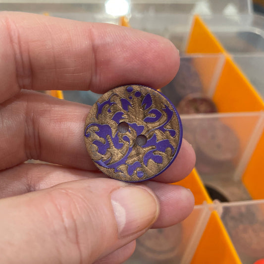 Hand holding a decorative button with a pattern, against a background of a button collection box.