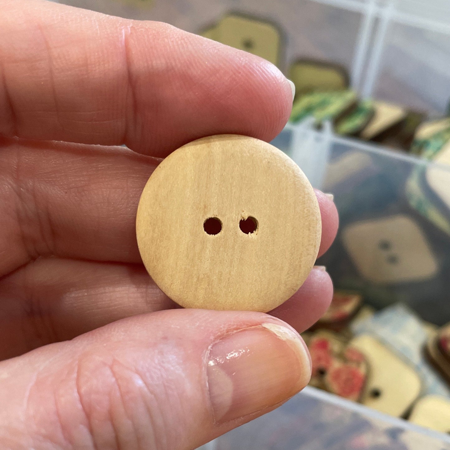 Hand holding a wooden button with a background of various buttons in containers.