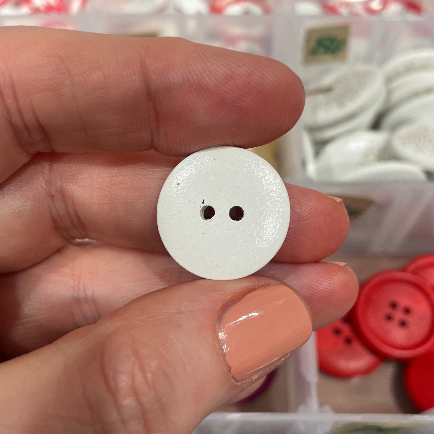 Hand holding a white button with a blurred container of buttons in the background