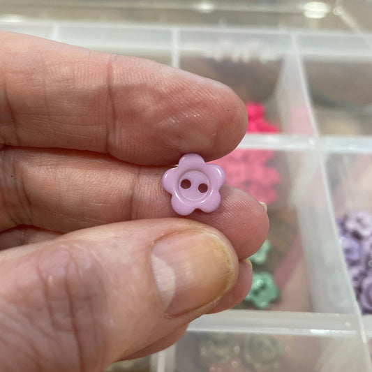 Hand holding a pink flower-shaped button with a container of buttons in the background