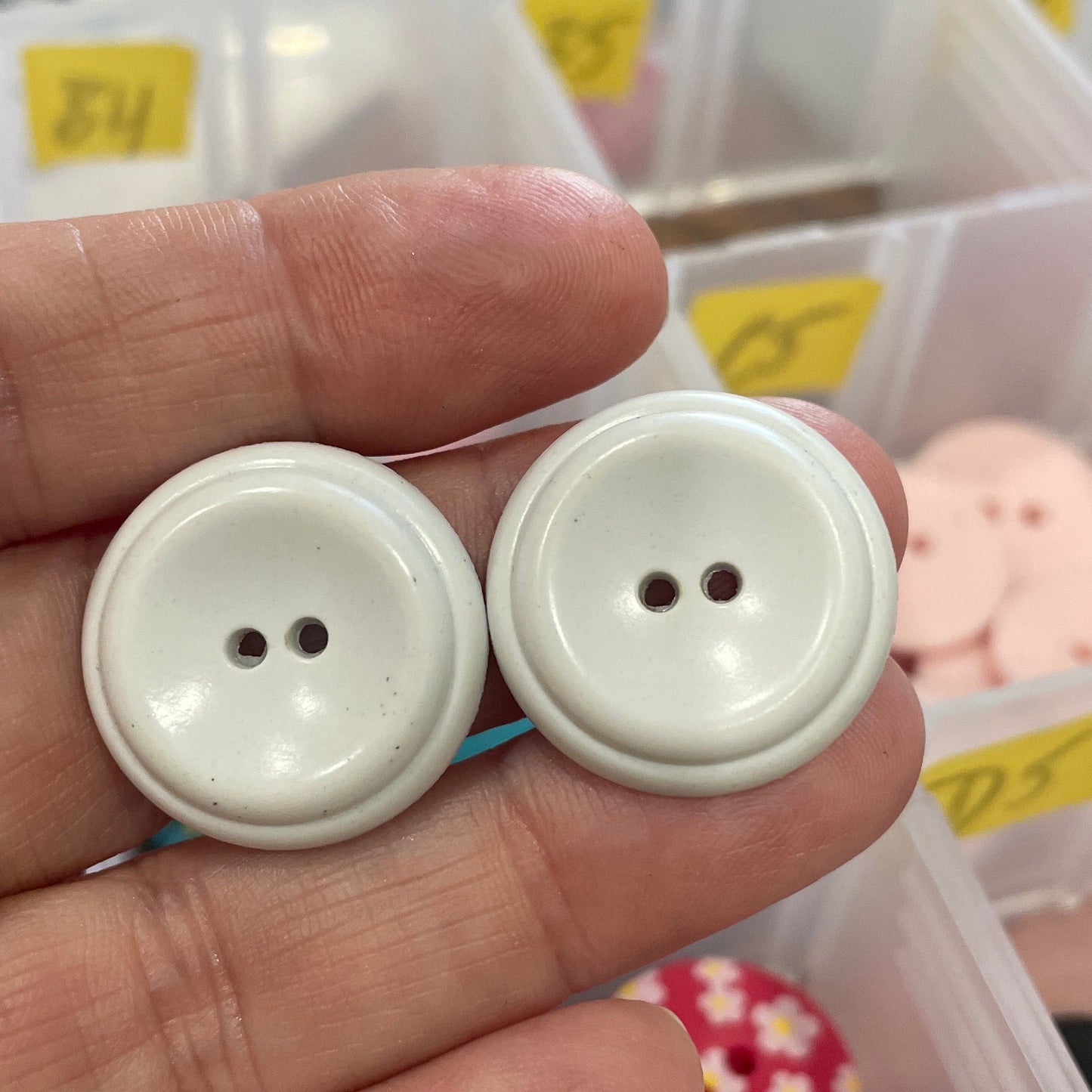 White buttons held between fingers with a container of buttons in the background