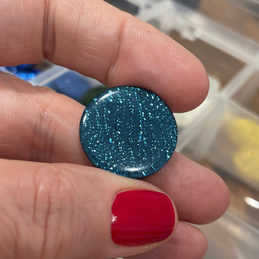Hand holding a blue glittery round object with a blurred background of buttons.
