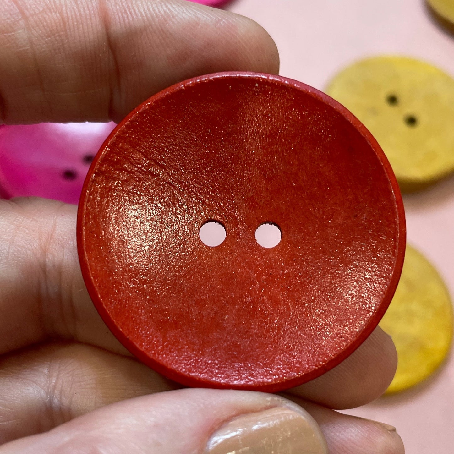 Hand holding a red button with two holes against a pink background with other buttons.