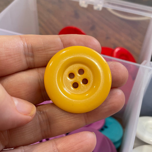 Hand holding a yellow button with a background of various buttons in a container.