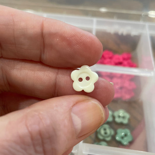 Small yellow flower-shaped button held between fingers with a container of similar earrings in the background.