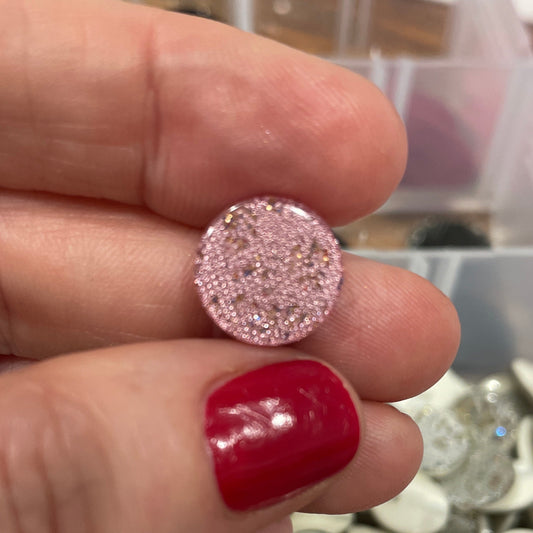 Pink glittery round earring held between fingers with a blurred background of buttons.
