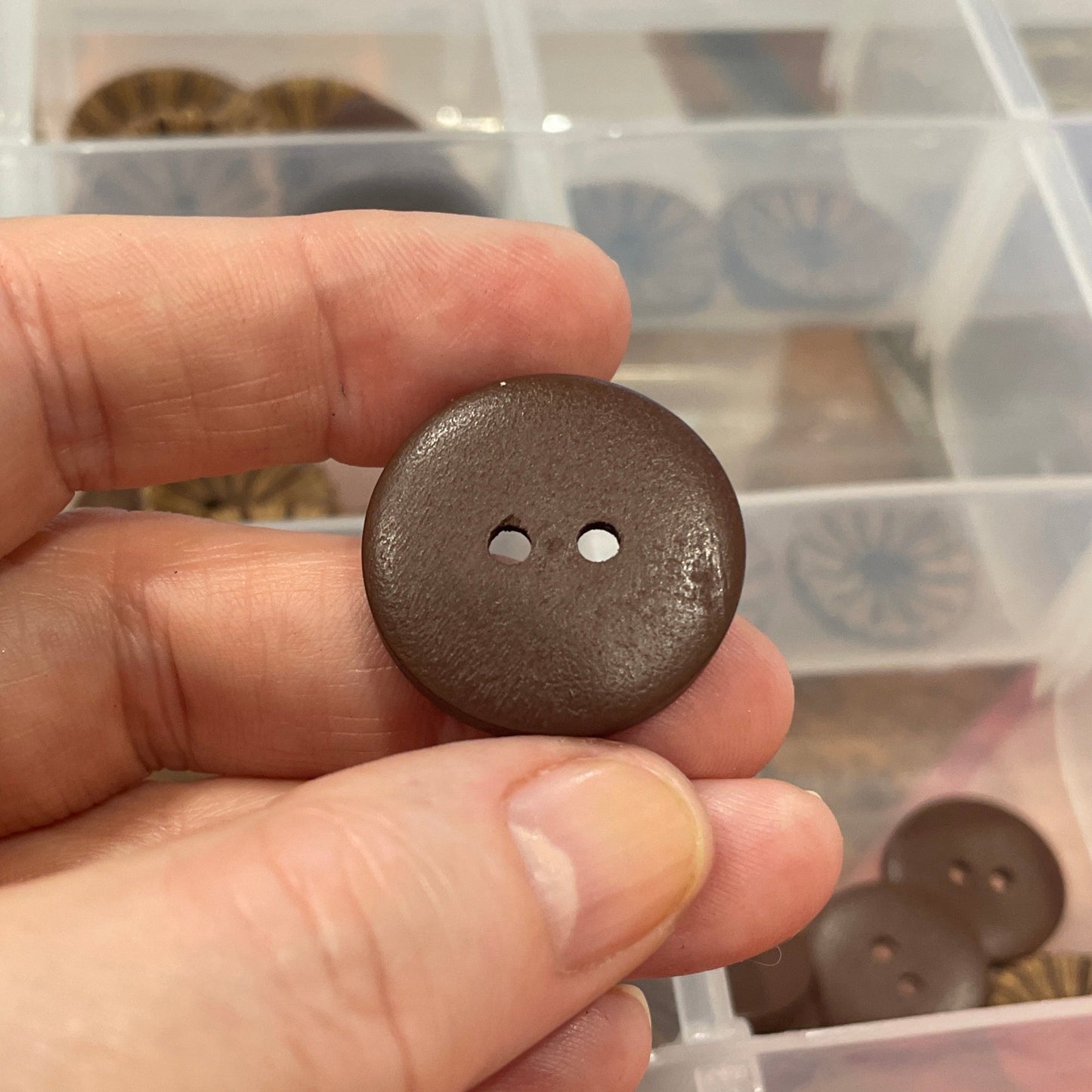 Hand holding a brown button with a clear container of buttons in the background
