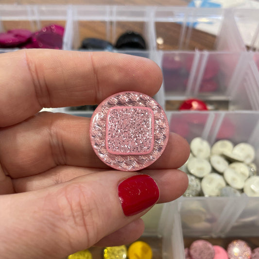 Hand holding a pink decorative button with a container of buttons in the background
