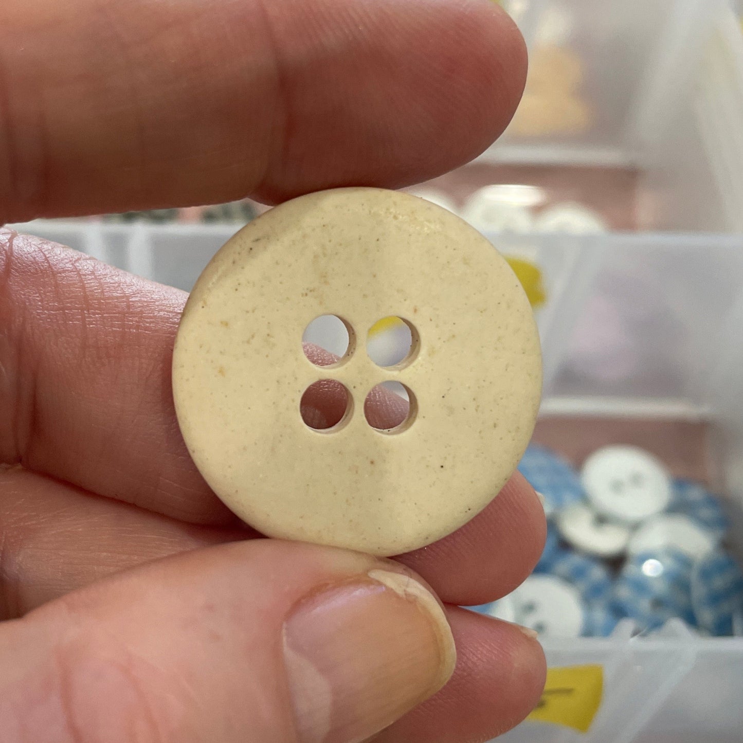 Hand holding a beige button with a clear container of buttons in the background