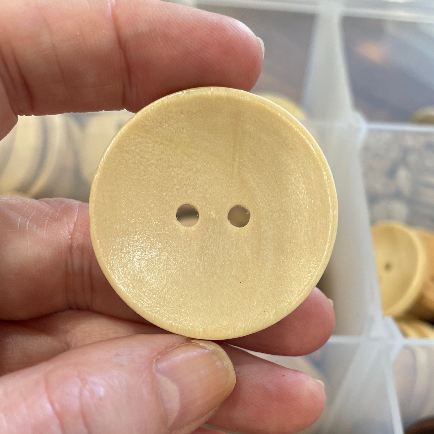 Hand holding a round beige button with a clear container of buttons in the background