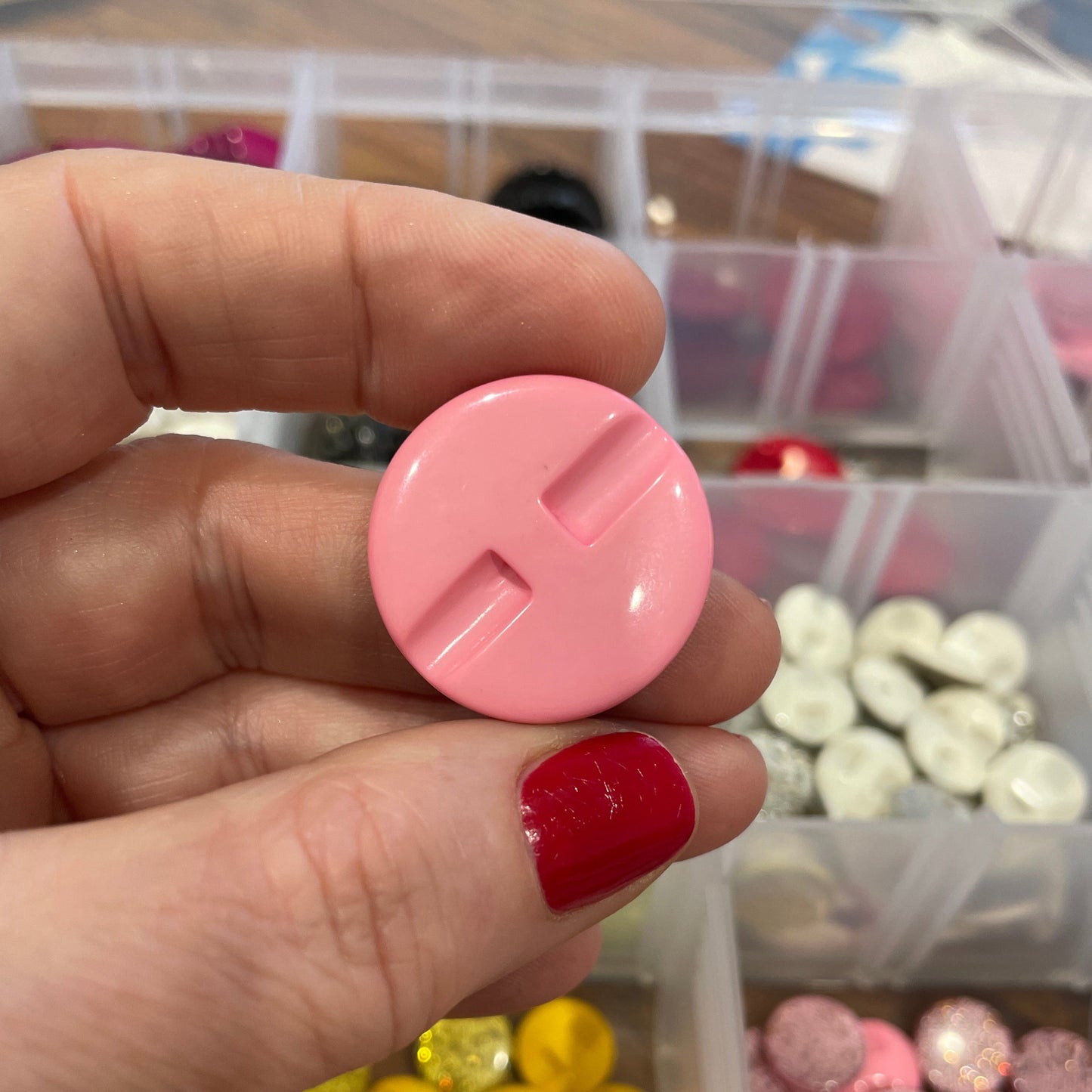 Hand holding a pink round object with a blurred background of colorful buttons in containers.