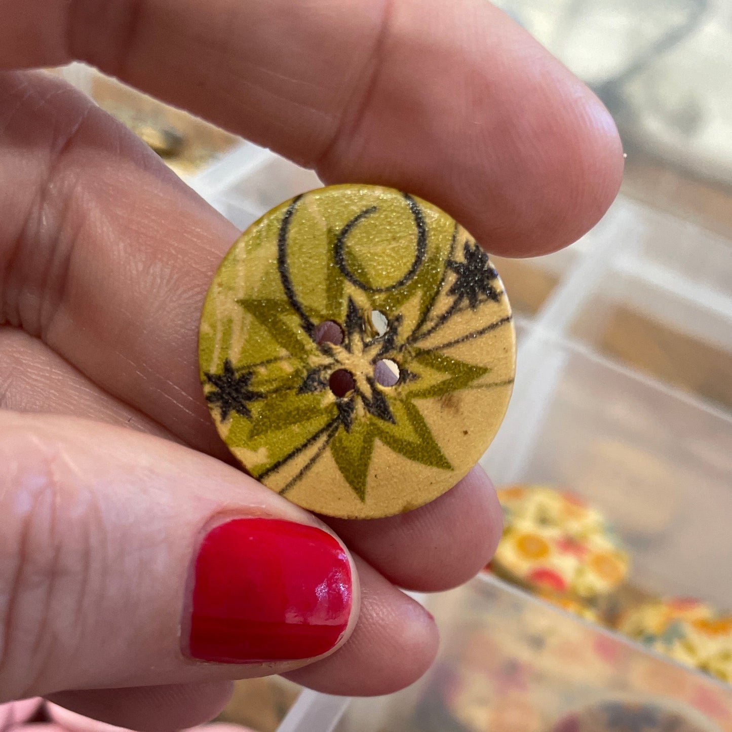 Hand holding a small round wooden button with floral design, in front of a container of various buttons.