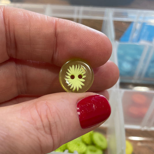 Hand holding a small gold button with a decorative center, against a background of button assortment.