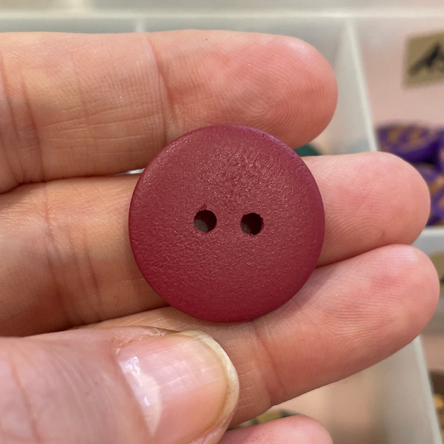 Maroon button held in a hand with a background of various buttons in a container.