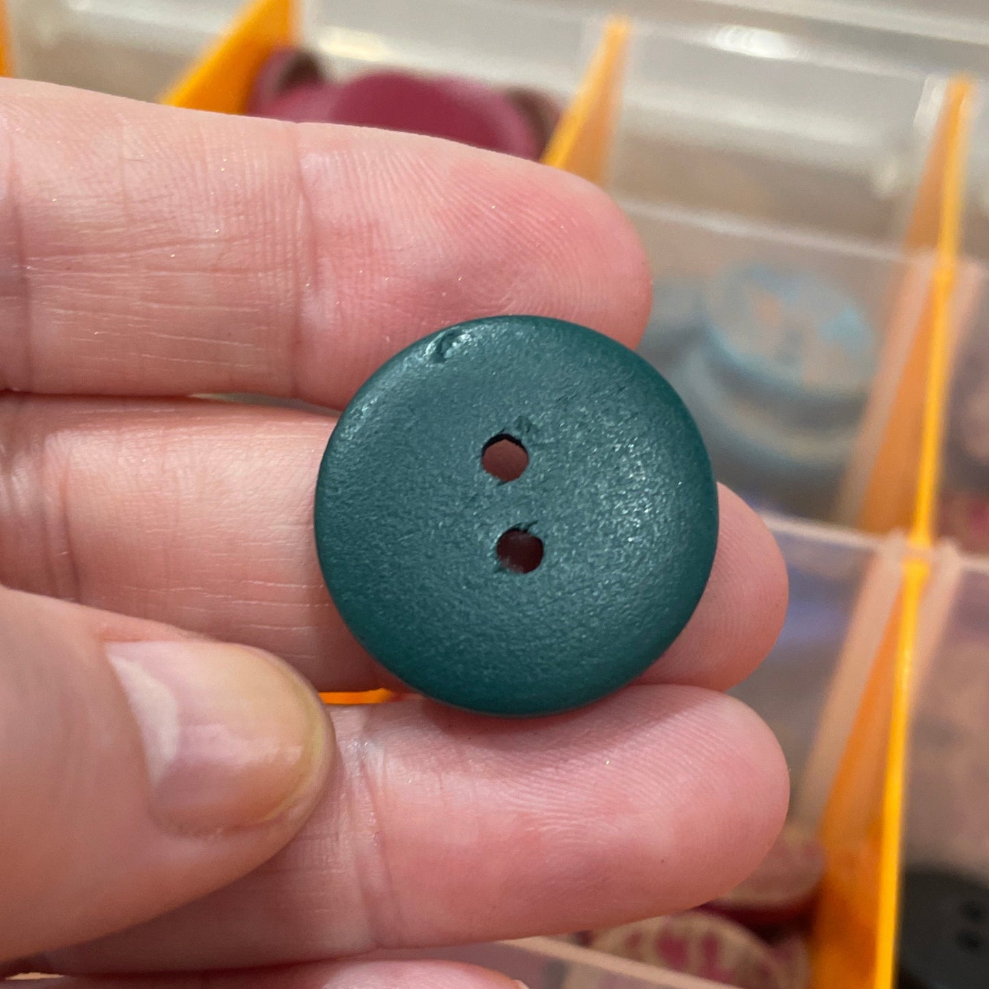 Hand holding a blue button with a clear container of buttons in the background