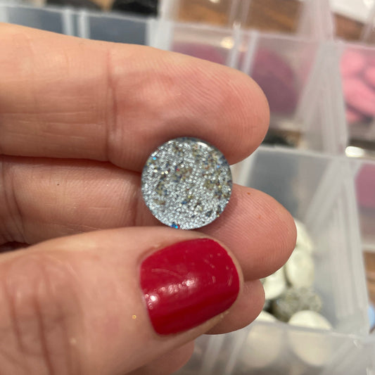Silver glittery round object held between fingers with a container of similar objects in the background.