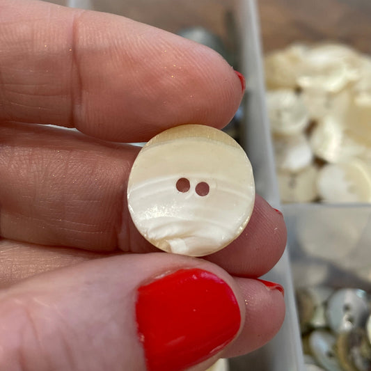 Hand holding a round beige button with two holes, with a container of buttons in the background.