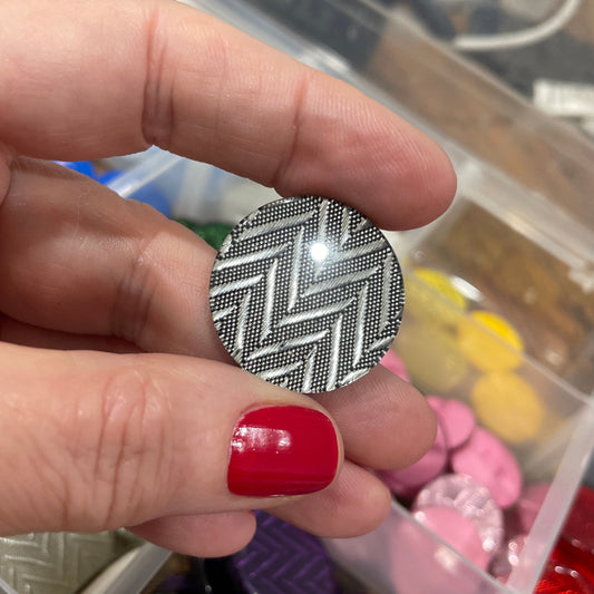 Hand holding a black and white patterned button with a container of colorful buttons in the background.