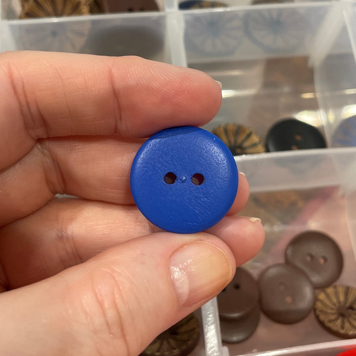 Hand holding a blue button with a clear container of assorted buttons in the background