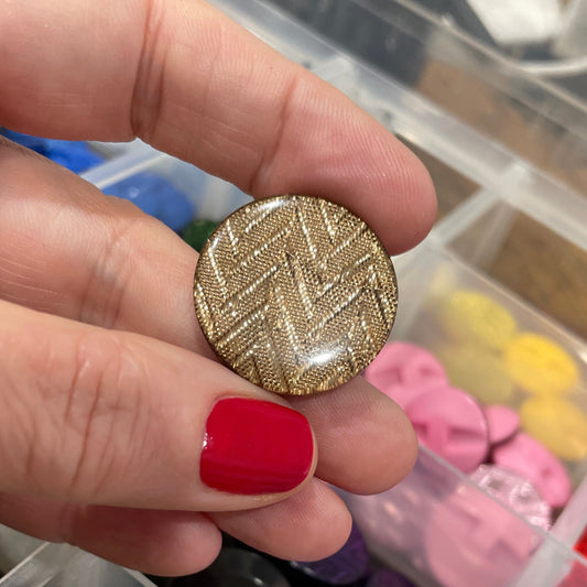Hand holding a gold textured button with a container of colorful buttons in the background