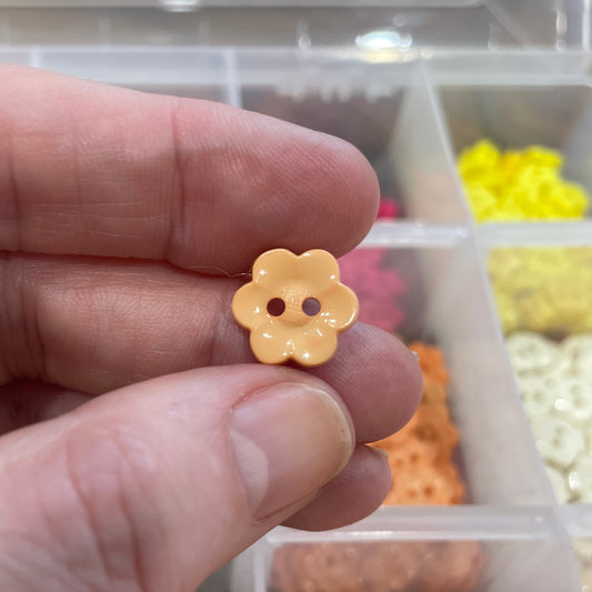 Hand holding a yellow flower-shaped button with a background of similar buttons in a container.