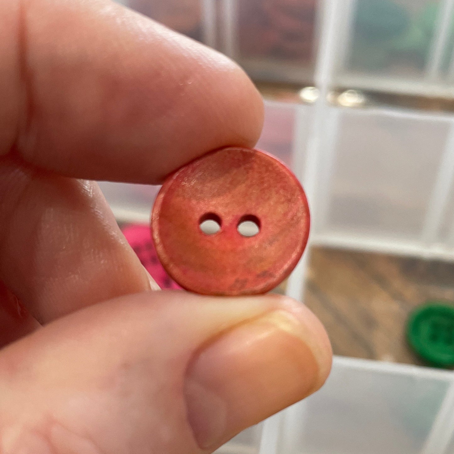 Hand holding a red button with two holes in front of a container of colorful buttons.