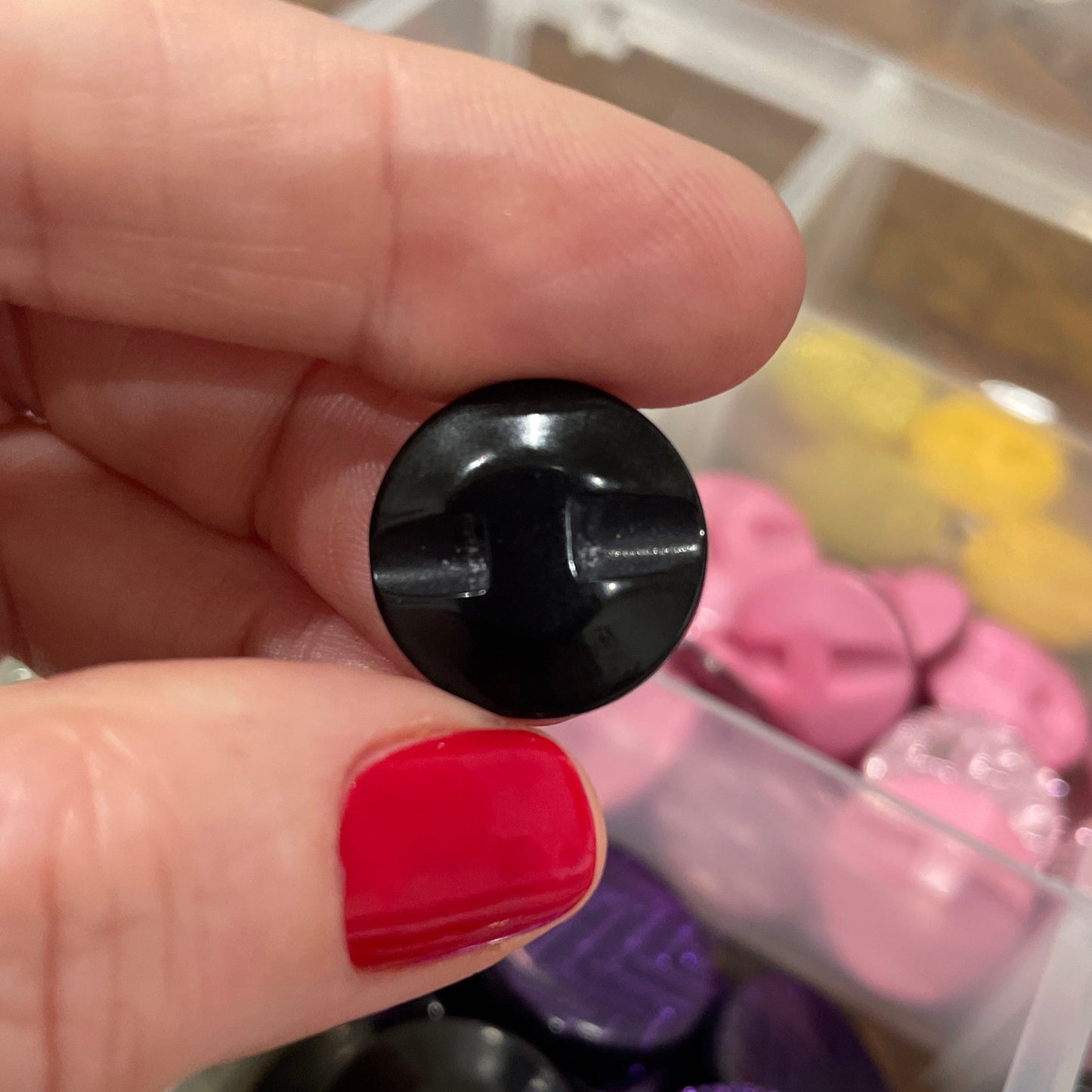 Black button held by a hand with red nail polish, against a background of colorful buttons in a container.