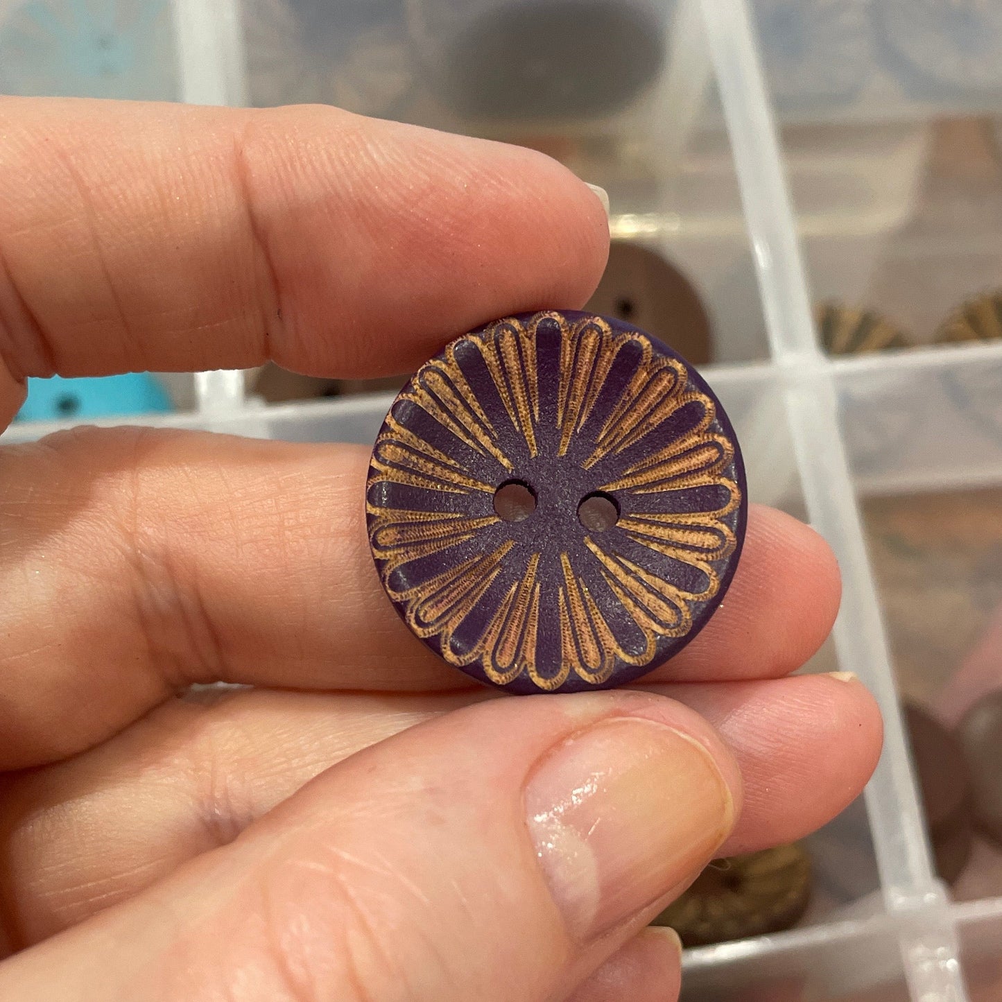 Hand holding a decorative button with a background of a button collection in a box.