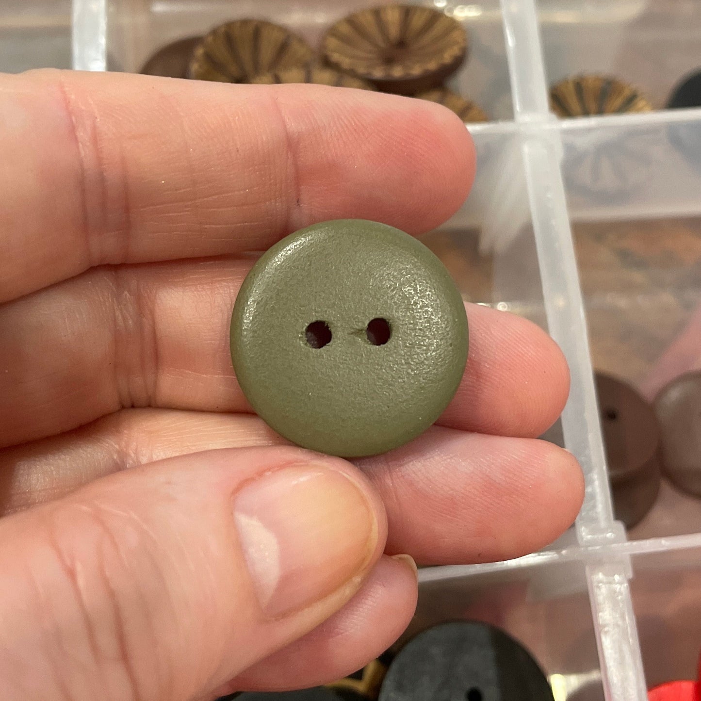Hand holding a green button with a clear container of assorted buttons in the background