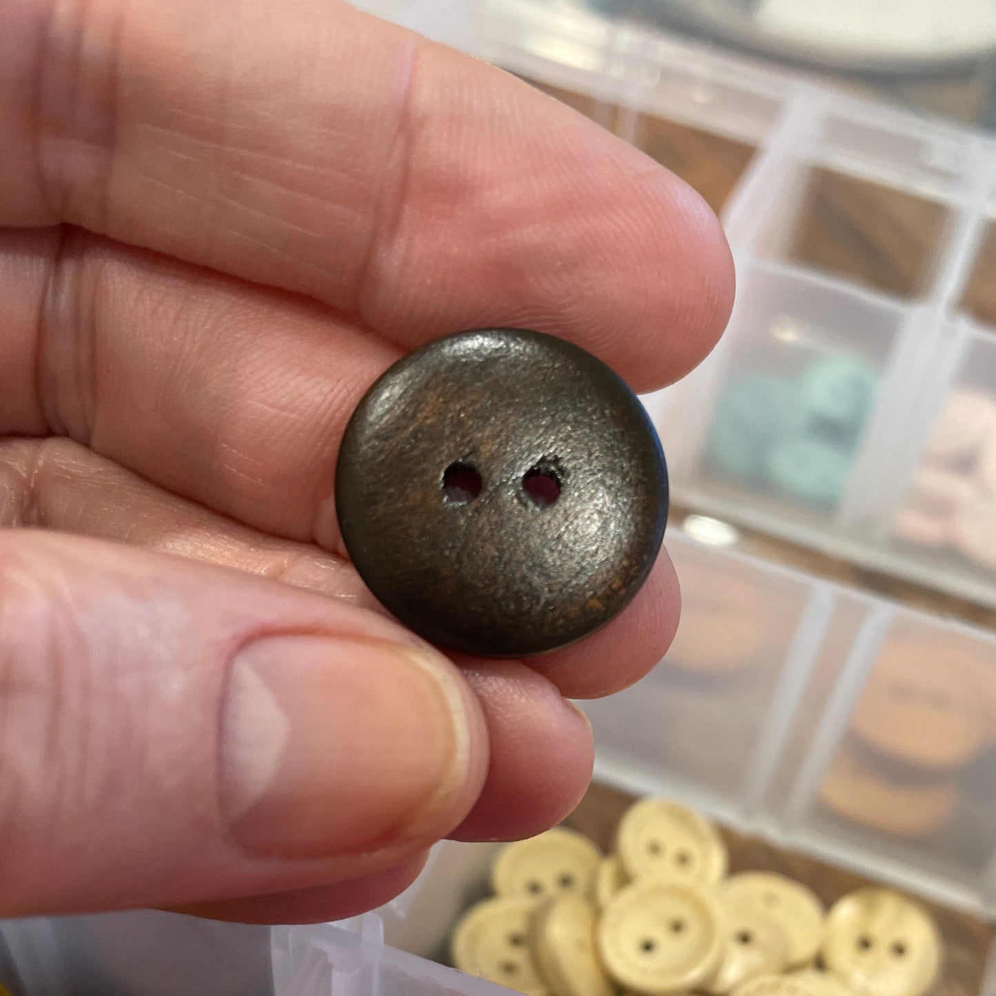 Hand holding a dark brown button with a background of colorful buttons in containers.