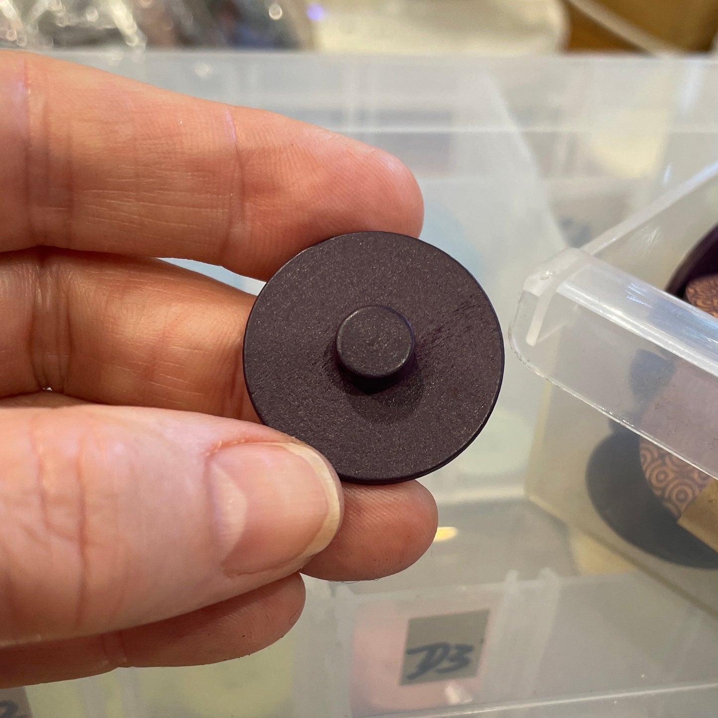 Hand holding a round brown button with a container of buttons in the background