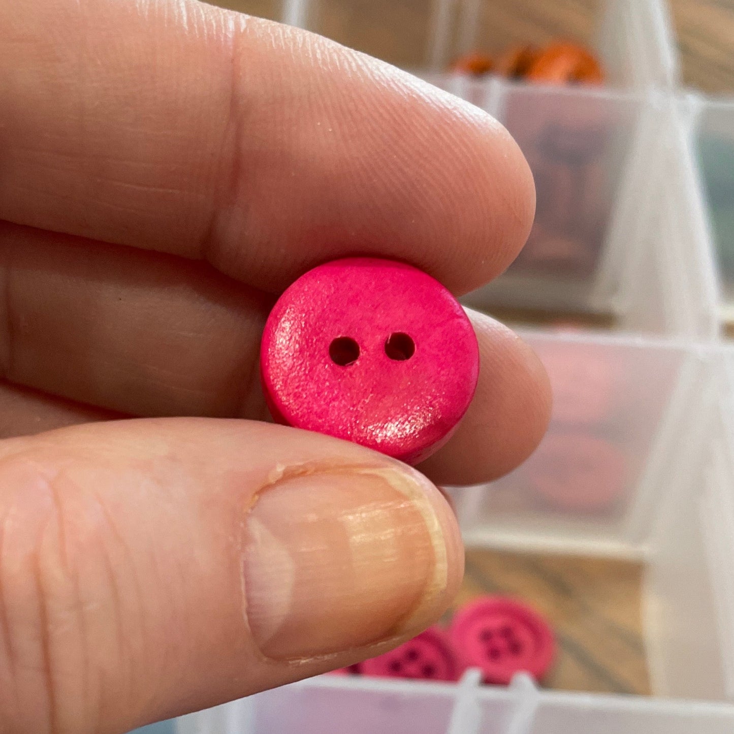 Hand holding a pink button with a container of assorted buttons in the background