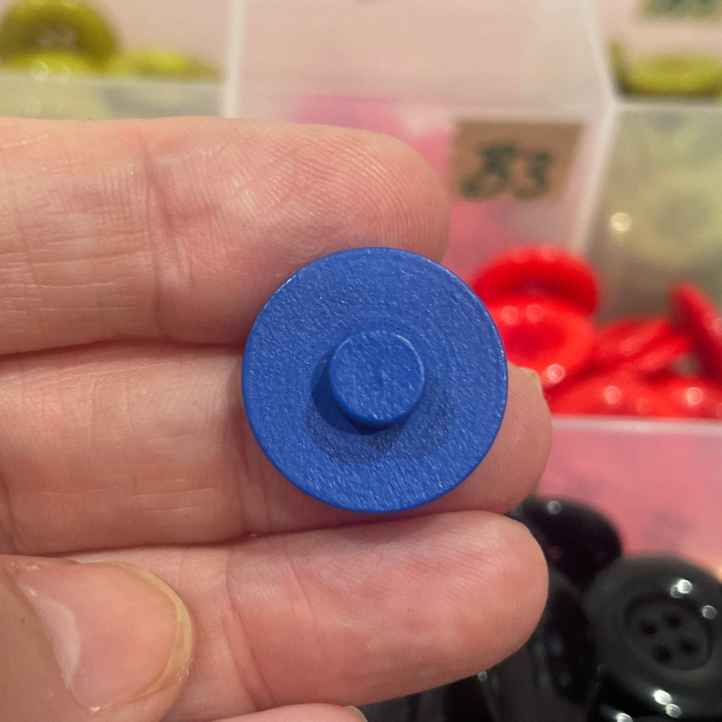 Hand holding a blue button with a background of assorted buttons in containers