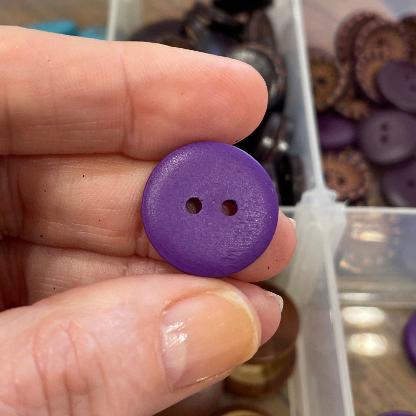 Hand holding a purple button with a container of various buttons in the background