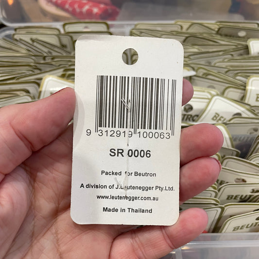 Hand holding a product tag with a barcode and text, with a container of similar products in the background.