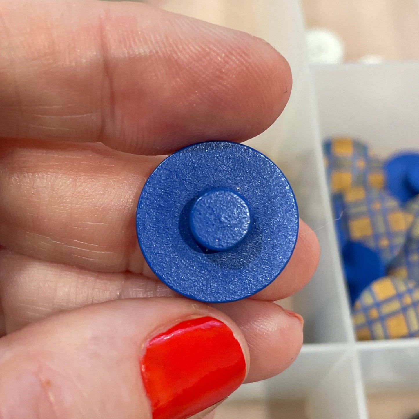 Hand holding a blue button with a container of buttons in the background