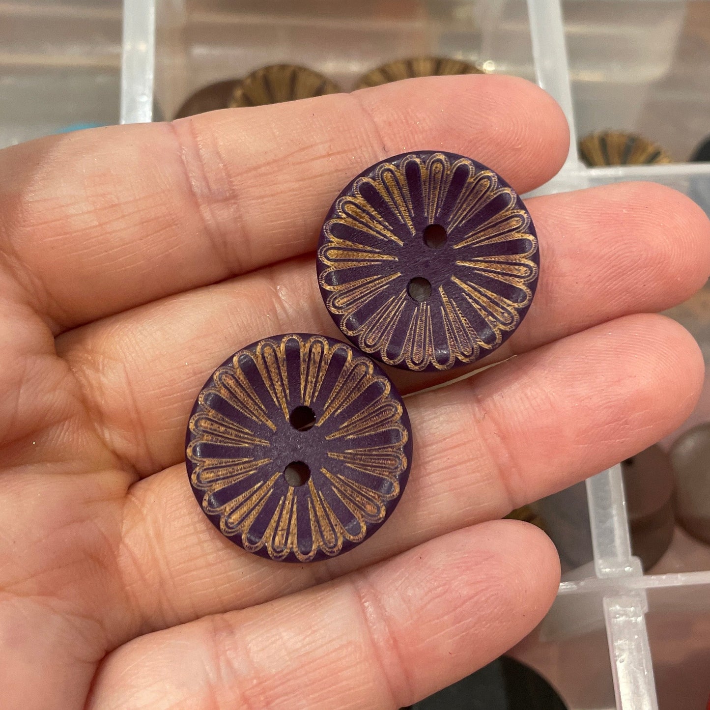 Hand holding two decorative buttons with a container of various buttons in the background