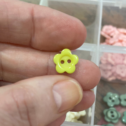 Hand holding a small yellow flower-shaped earring with a display of various earrings in the background.