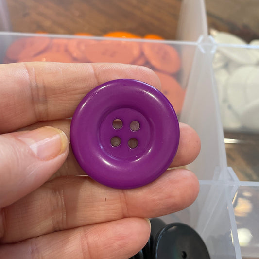 Hand holding a purple button with a clear container of buttons in the background