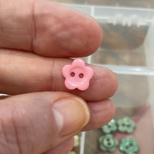 Pink flower-shaped button held between fingers with a container of similar buttons in the background.