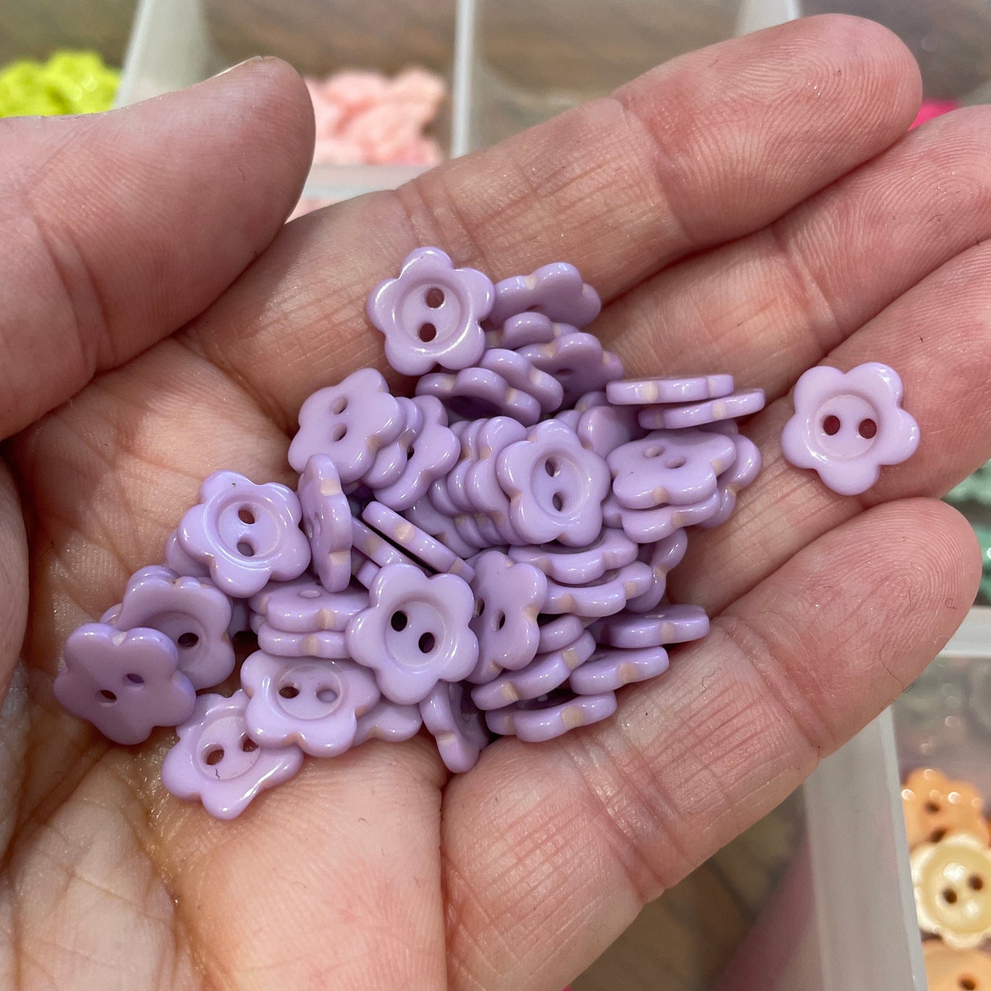 Hand holding small purple flower-shaped beads with a blurred background of more beads.