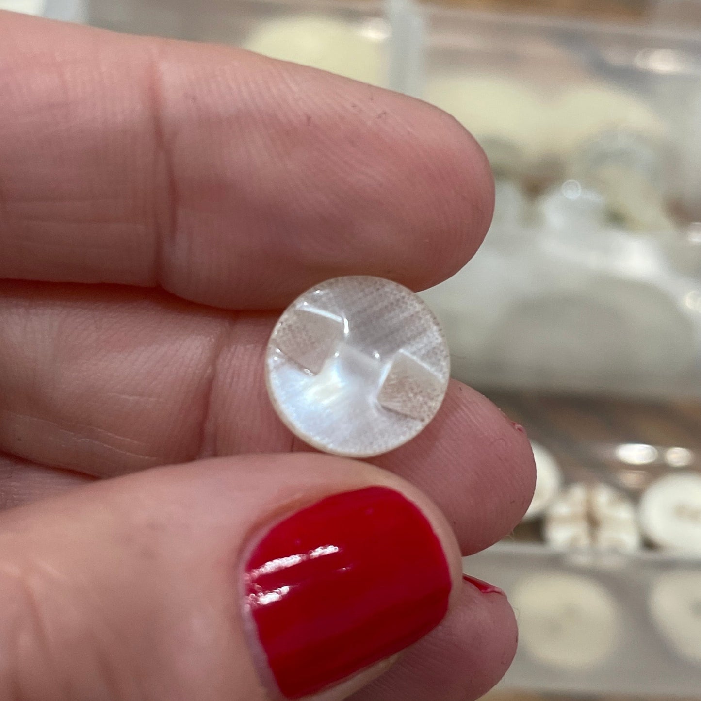 Hand holding a round, silver-colored earring with a clear container of earrings in the background.