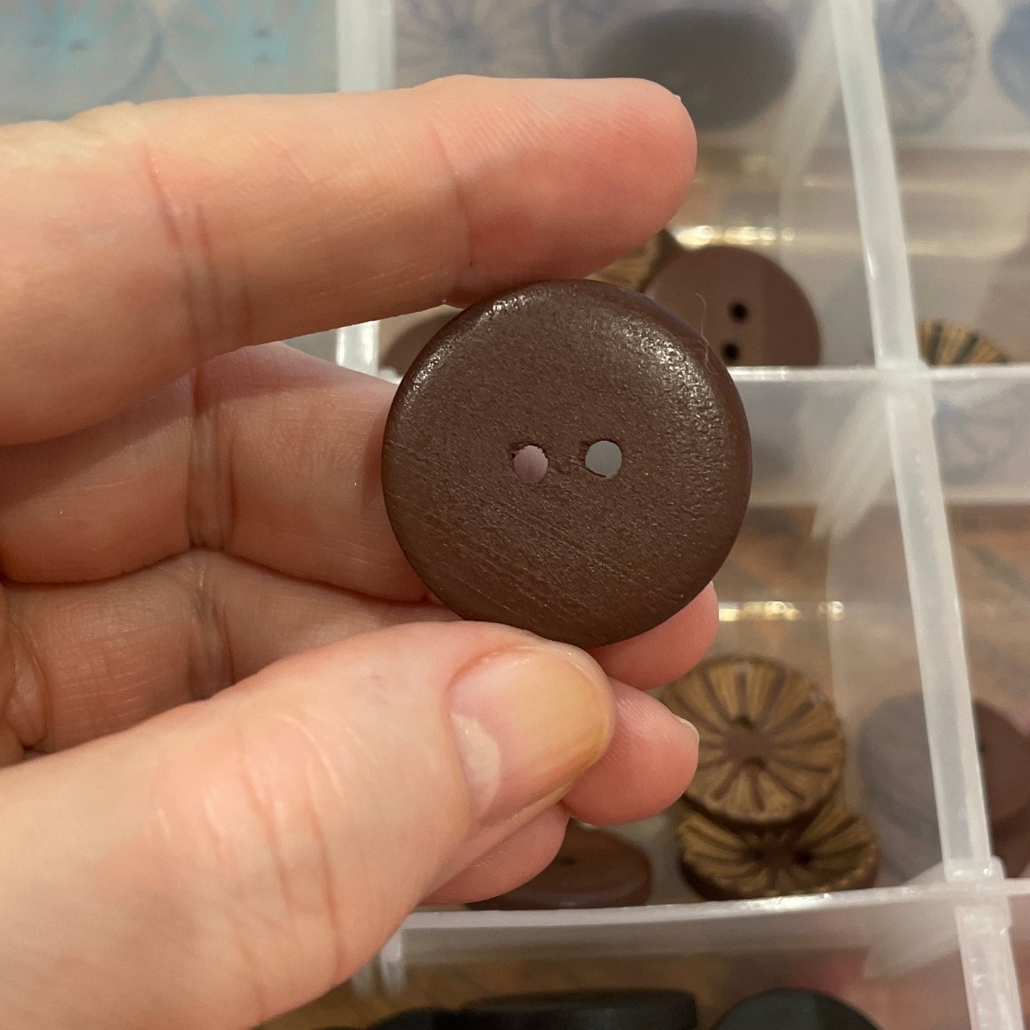 Hand holding a brown button above a container of assorted buttons.