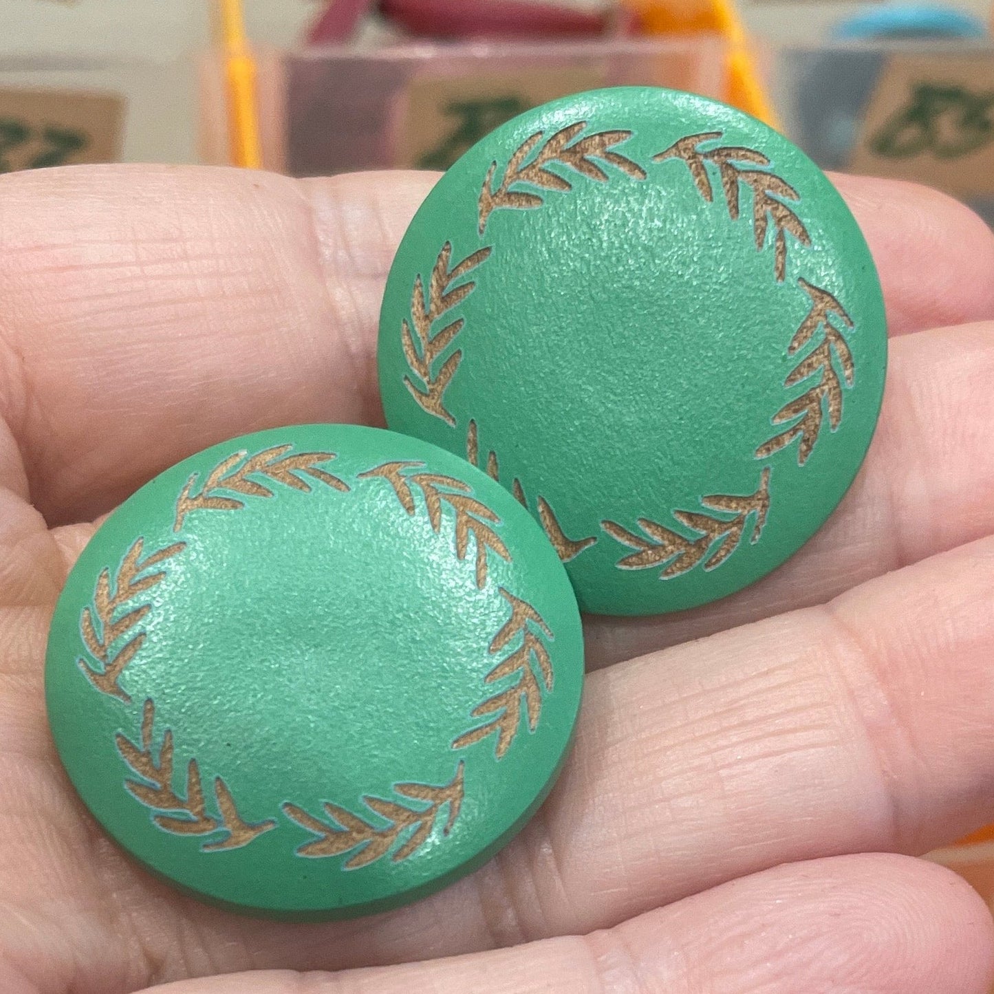 Two green buttons with gold patterns held in front of a container of assorted buttons.